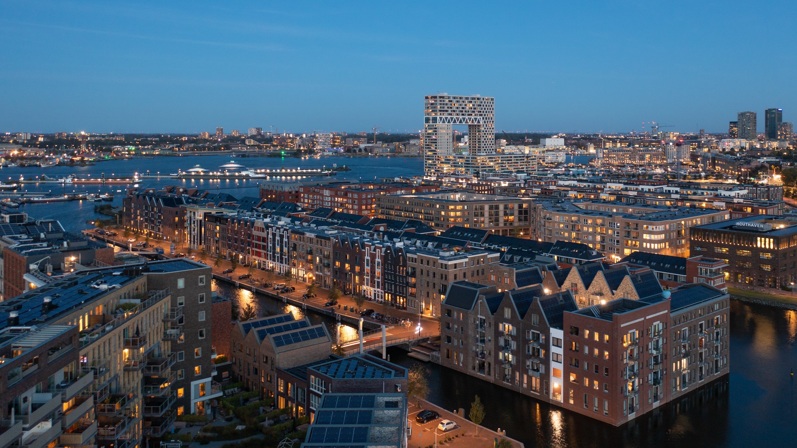 Luchtfoto bij avondschemering van de kadewoningen met pakhuisgevels op het Wiborg-eiland in de Amsterdamse Houthavens, aan het water.